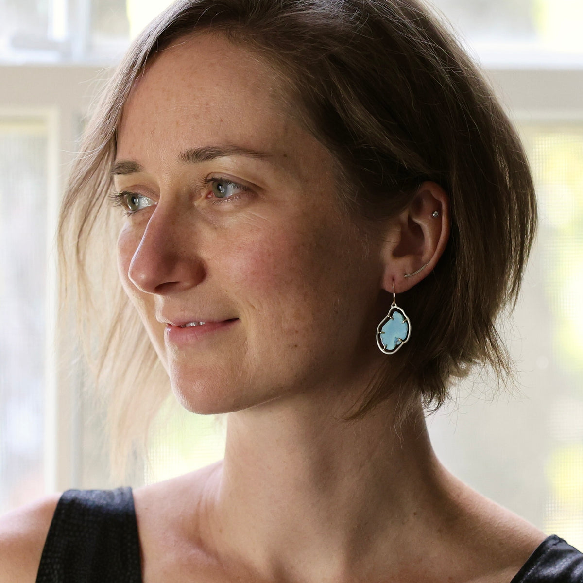 A woman with short light brown hair is shown in profile wearing a dangling earring with a light blue oval stone in a silver-toned setting.