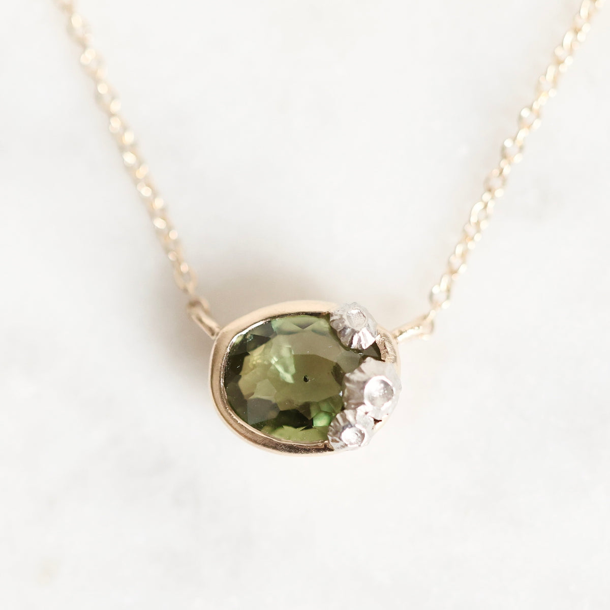 A close-up of a gold necklace with a pendant featuring a faceted olive green gemstone and small clear stones, shown on a light marble-like background.