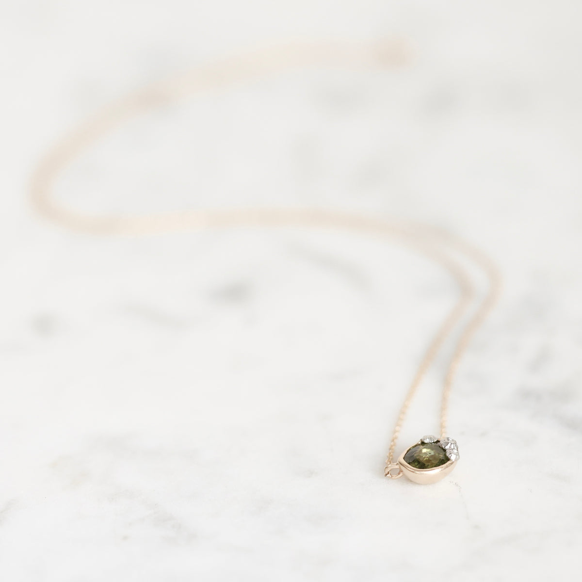A delicate rose gold necklace with a small oval green gemstone pendant, resting on a white marble surface.