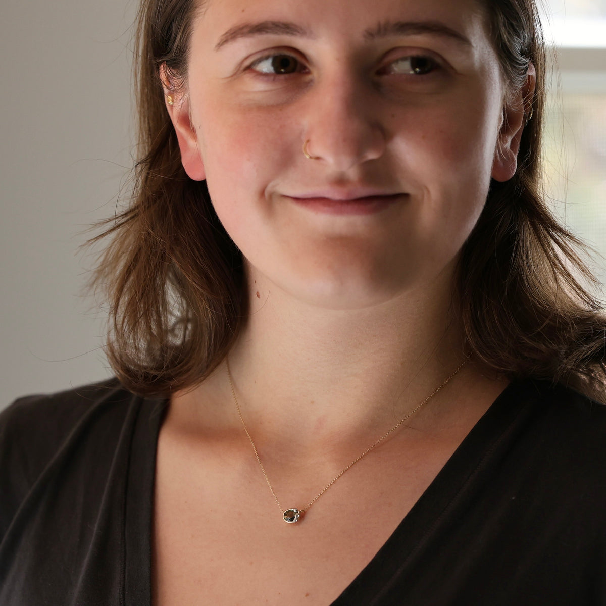 A woman with shoulder-length brown hair wearing a delicate gold chain necklace with a small round dark bead pendant, shown in a close-up portrait.