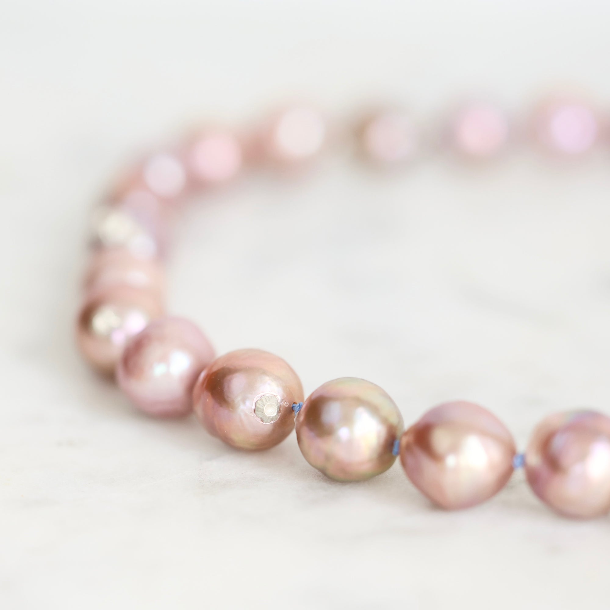 A close-up of a beaded strand bracelet made of iridescent pink baroque pearls strung on a thin blue cord, resting on a light marble surface.