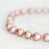 A close-up of a beaded strand bracelet made of iridescent pink baroque pearls strung on a thin blue cord, resting on a light marble surface.