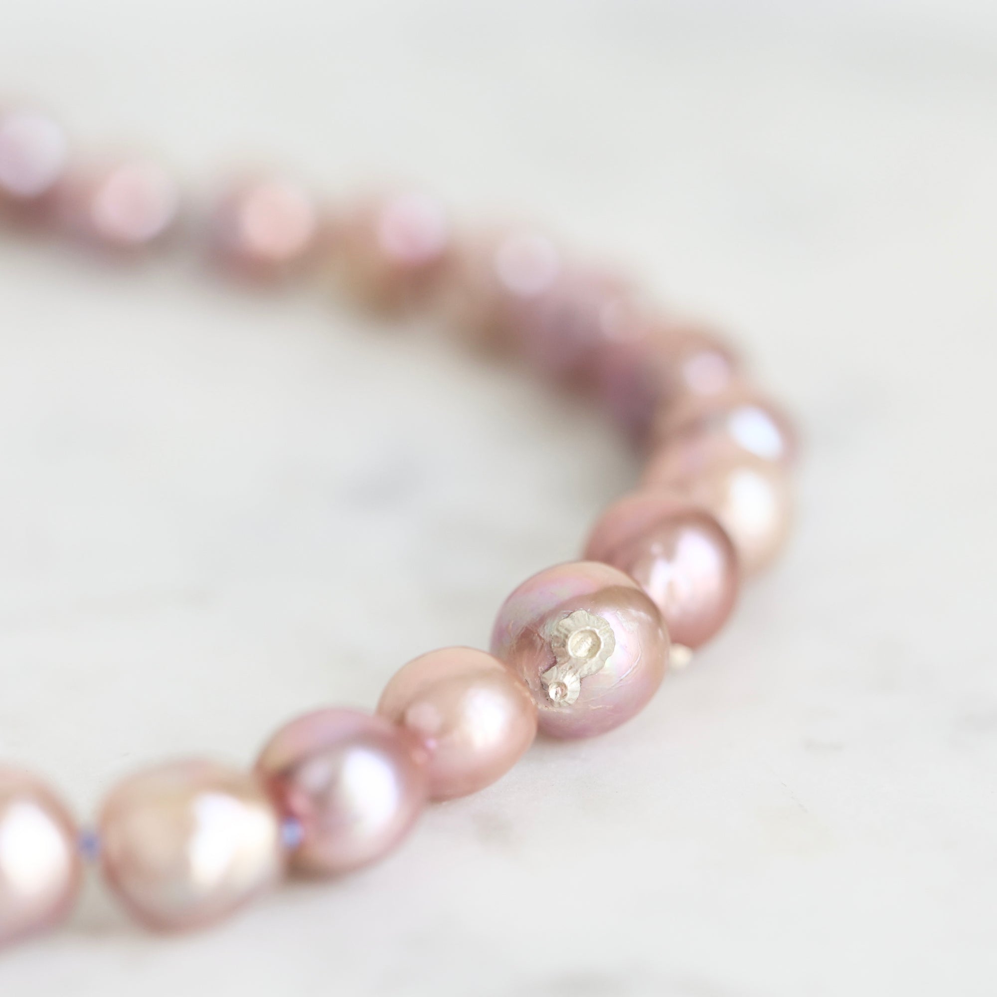 A close-up of a beaded bracelet made of irregular, lustrous pinkish pearl-like beads with a small gold charm, resting on a light marble surface.