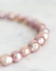 A close-up of a beaded bracelet made of irregular, lustrous pinkish pearl-like beads with a small gold charm, resting on a light marble surface.
