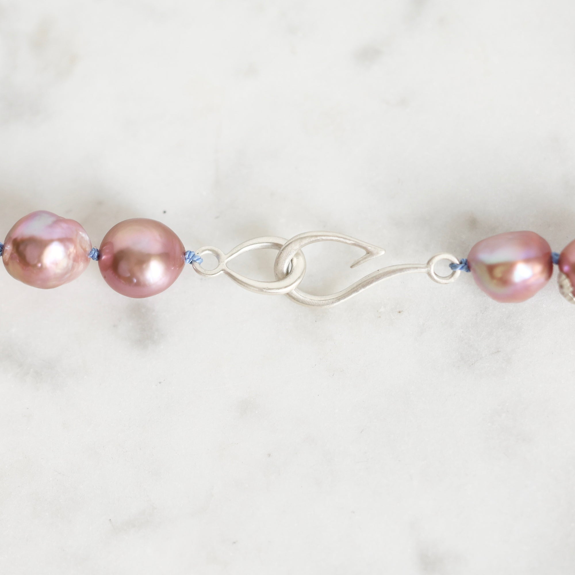 A close-up of a jewelry piece featuring iridescent pink baroque pearls threaded on a blue cord with a polished silver clasp, set against a light marble background.