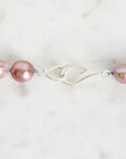 A close-up of a jewelry piece featuring iridescent pink baroque pearls threaded on a blue cord with a polished silver clasp, set against a light marble background.