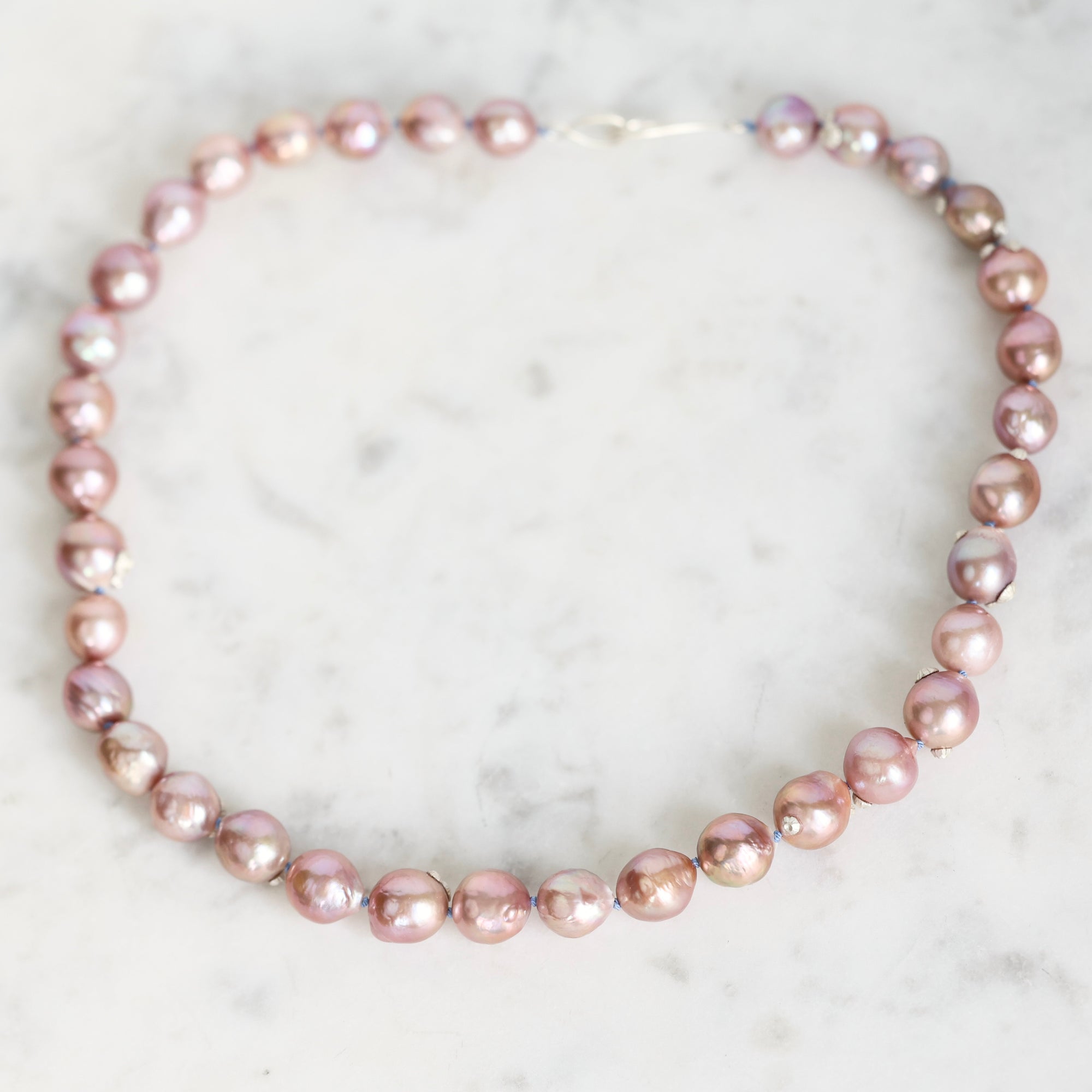 A strand of irregular baroque pearls in a dusty pink color, arranged in a circle on a white marble surface.