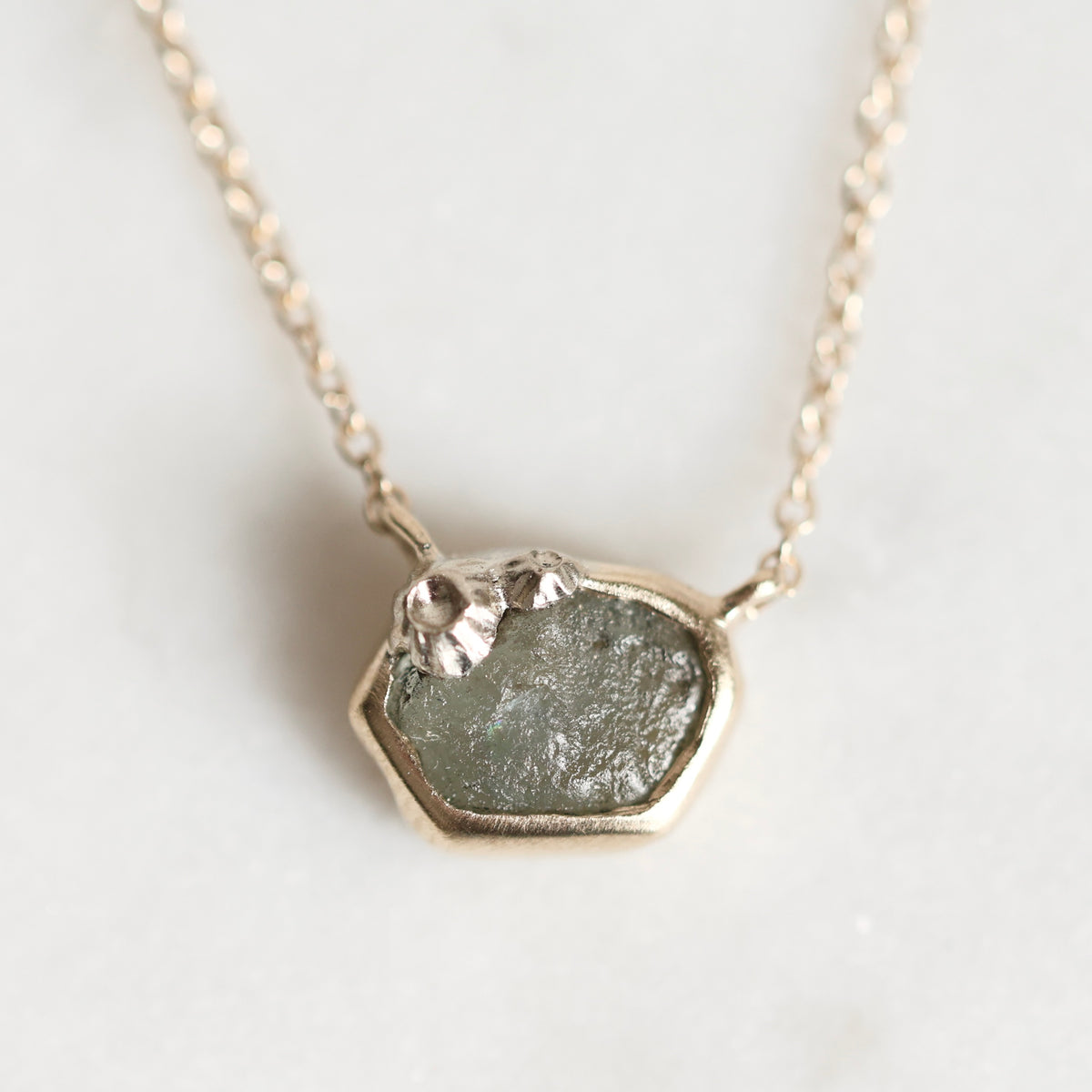 A close-up shot of a delicate gold chain necklace featuring a unique, irregular-shaped pendant. The pendant is set with a textured, natural green stone and decorated with small, sculpted gold leaf pieces on top, resting on a light, neutral background.
