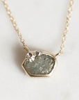 A close-up shot of a delicate gold chain necklace featuring a unique, irregular-shaped pendant. The pendant is set with a textured, natural green stone and decorated with small, sculpted gold leaf pieces on top, resting on a light, neutral background.