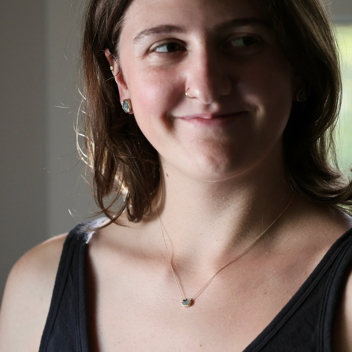 A woman with brown hair wearing a delicate gold chain necklace with a small heart-shaped stone and matching stud earrings, shown in a close-up portrait.