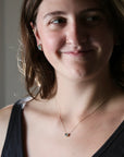 A woman with brown hair wearing a delicate gold chain necklace with a small heart-shaped stone and matching stud earrings, shown in a close-up portrait.