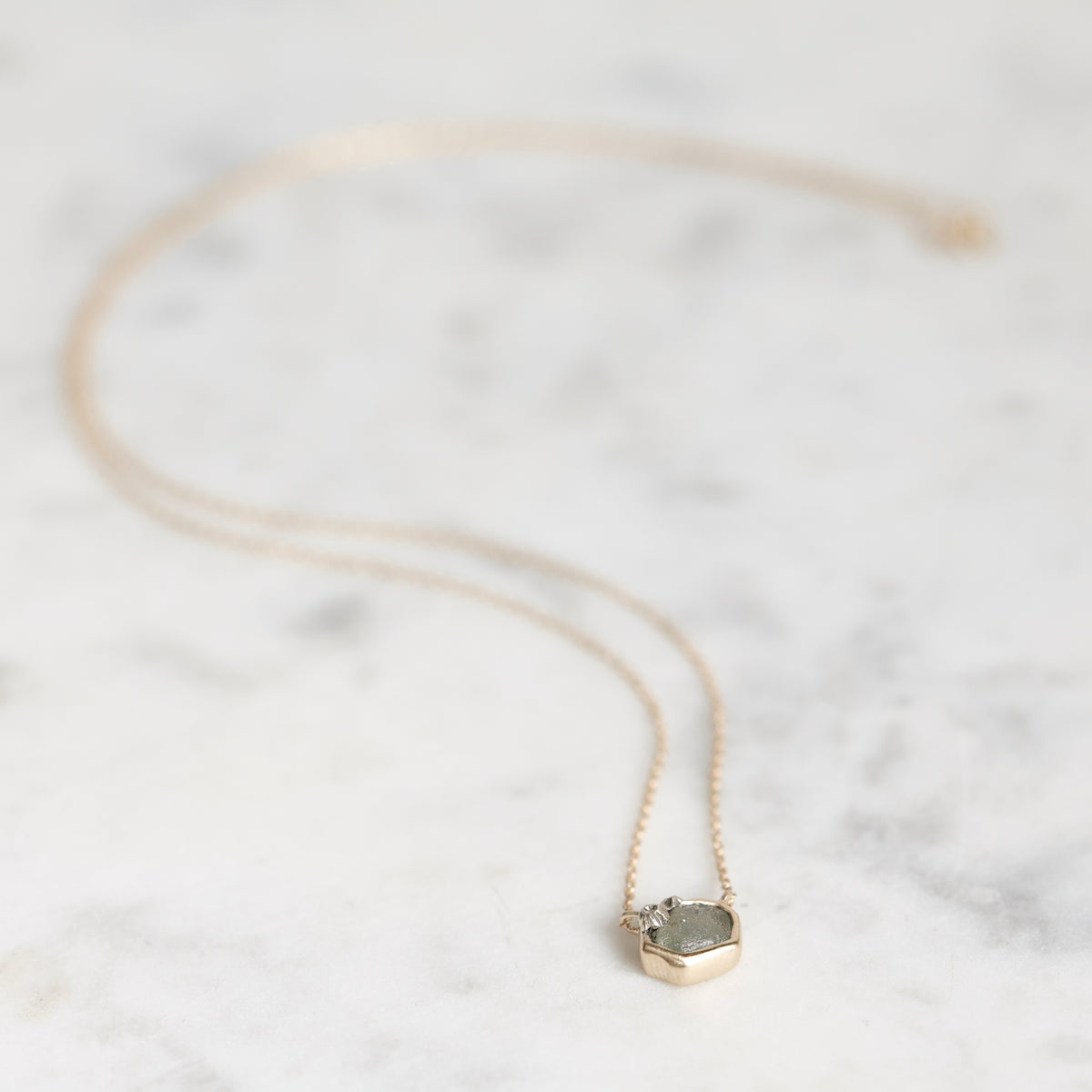 A delicate gold chain necklace with a small hexagonal pendant featuring a green stone and tiny clear accent stones, displayed on a light marble surface.