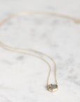 A delicate gold chain necklace with a small hexagonal pendant featuring a green stone and tiny clear accent stones, displayed on a light marble surface.