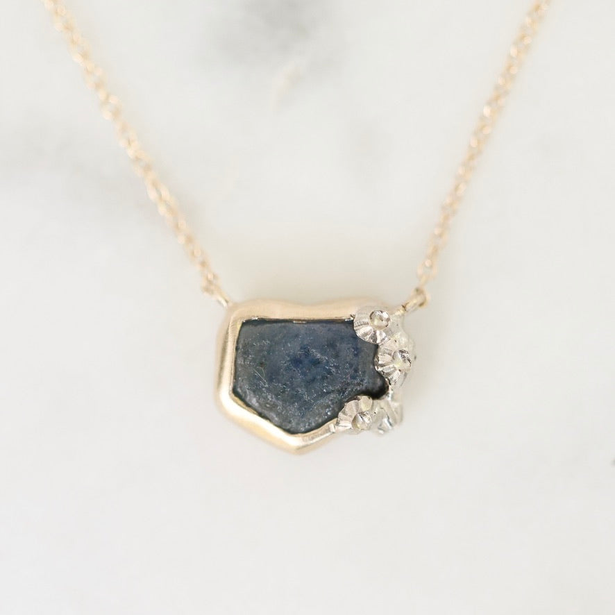 A gold necklace with a raw, irregular dark blue gemstone pendant and small clear crystals on one corner, displayed on a light marble surface.