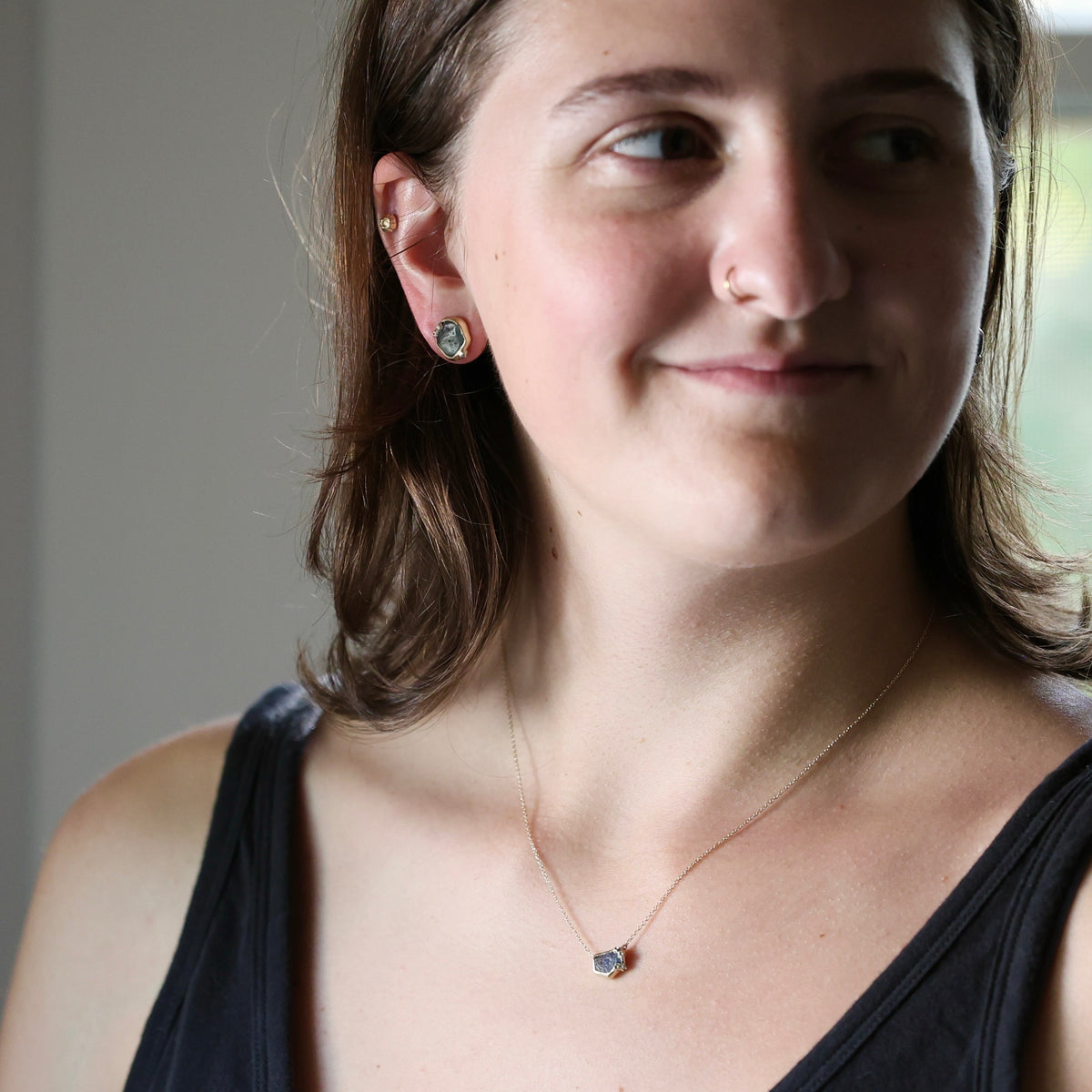 A close-up lifestyle shot of a woman wearing a delicate rose gold necklace with a small dark stone pendant and matching stud earrings.