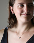 A close-up lifestyle shot of a woman wearing a delicate rose gold necklace with a small dark stone pendant and matching stud earrings.