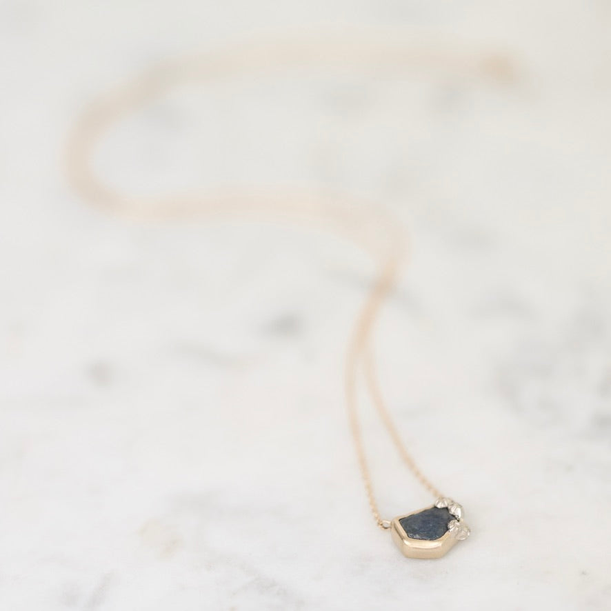 A delicate gold necklace with a raw, irregular dark gray stone pendant, resting on a light marble surface.