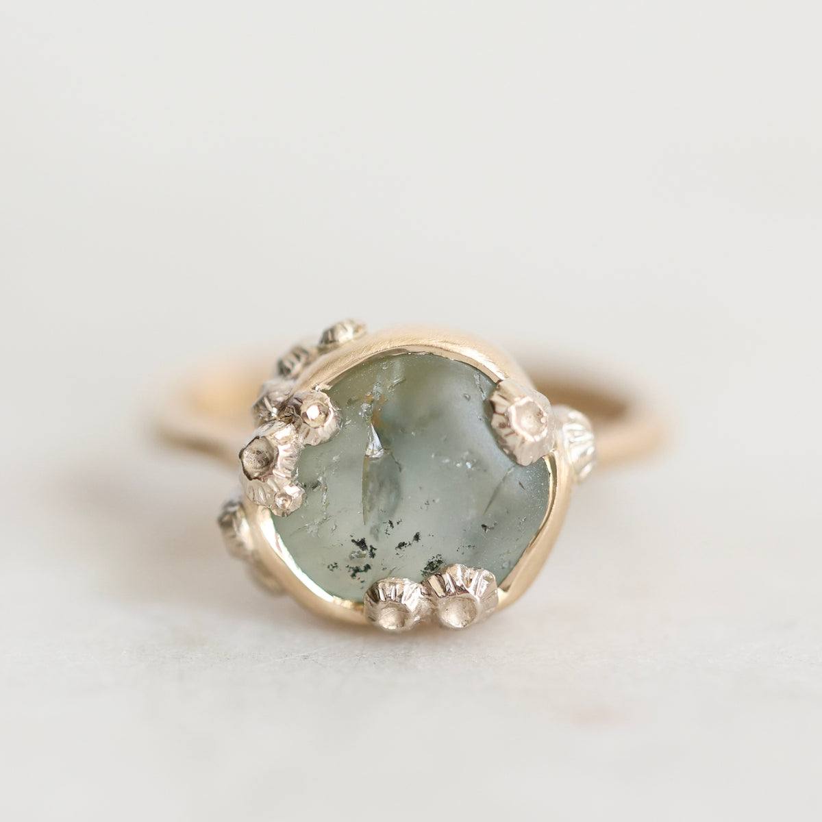 A close-up shot of a ring featuring a round, pale green stone with dark inclusions, surrounded by a gold-toned bezel and small silver-toned accents, set against a light off-white background.