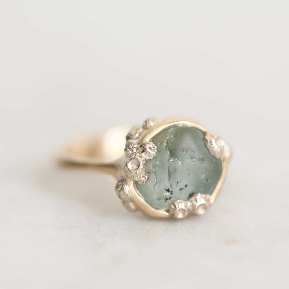 A close-up shot of a gold ring featuring a large, irregular pale green stone with tiny clear accent stones around its edge, set against a soft, light gray background.