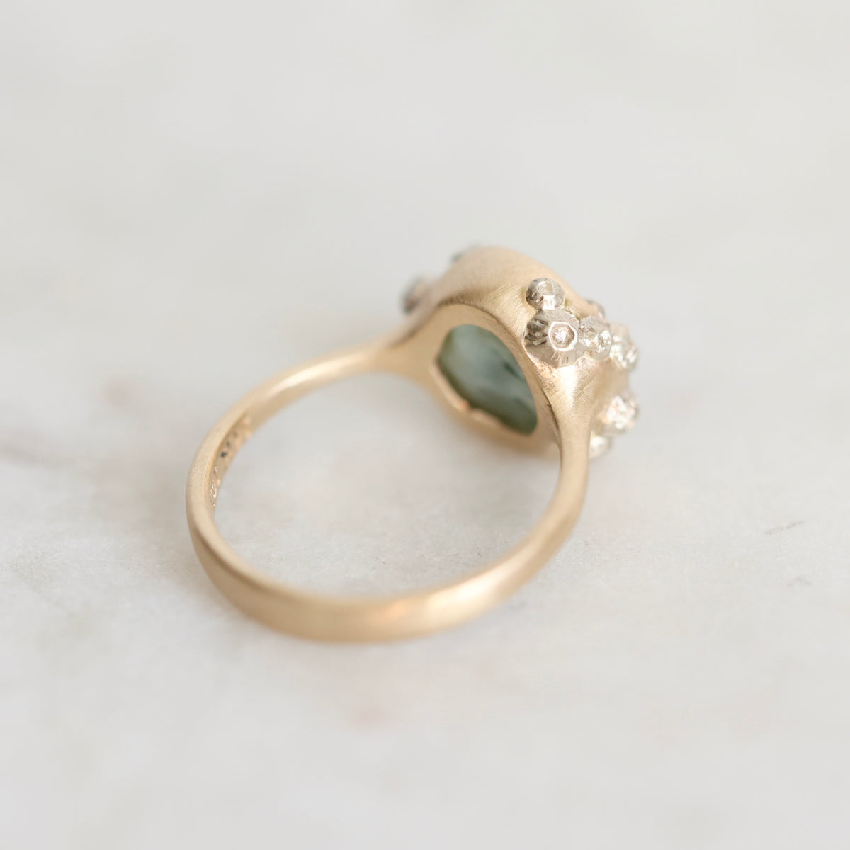 A gold ring with a textured, organic-shaped setting featuring a light green heart-shaped stone and small clear accent stones, photographed on a light marble surface.