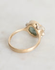 A gold ring with a textured, organic-shaped setting featuring a light green heart-shaped stone and small clear accent stones, photographed on a light marble surface.