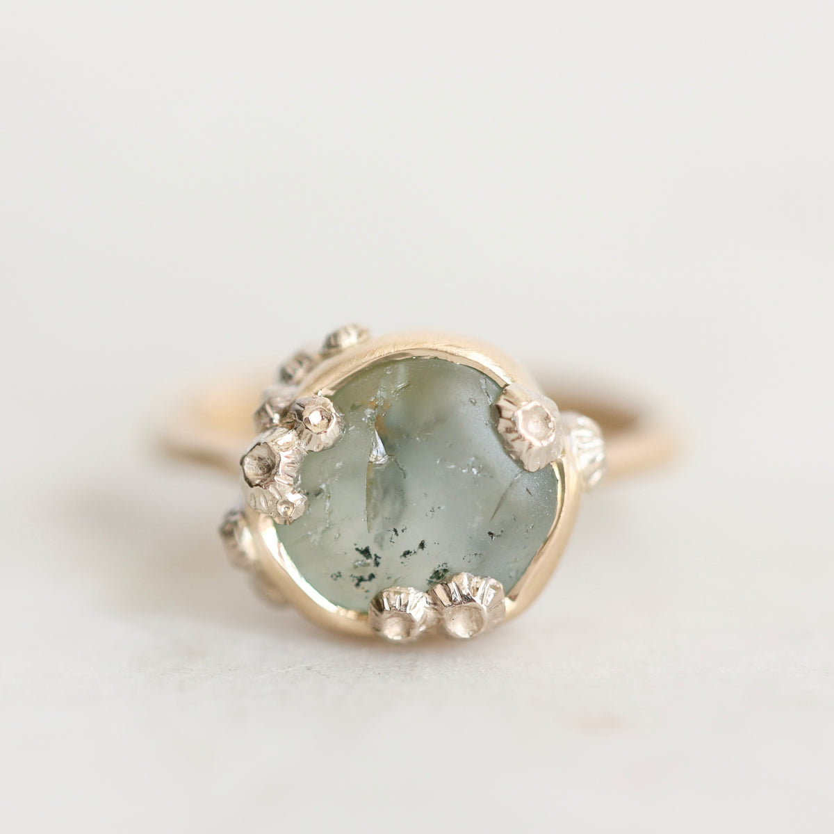 A close-up shot of a ring featuring a round, pale greenish-gray stone with dark inclusions, surrounded by small silver flower-like accents and a gold band, set against a light off-white background.