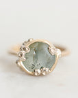 A close-up shot of a ring featuring a round, pale greenish-gray stone with dark inclusions, surrounded by small silver flower-like accents and a gold band, set against a light off-white background.