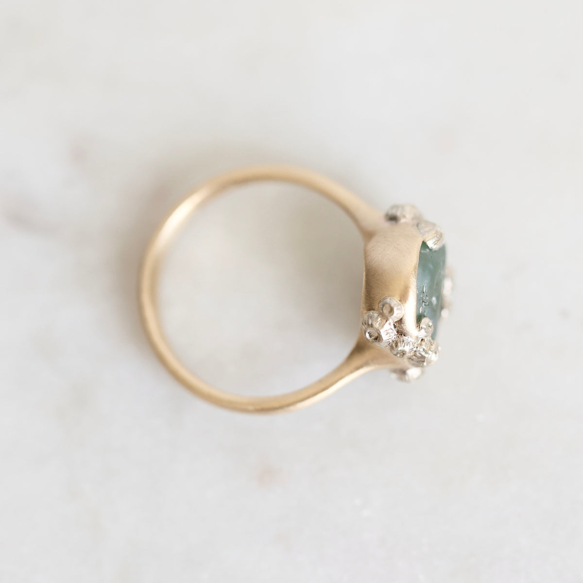 A gold ring with a green rectangular gemstone and small clear stones, photographed on a light marble surface.