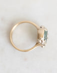 A gold ring with a green rectangular gemstone and small clear stones, photographed on a light marble surface.