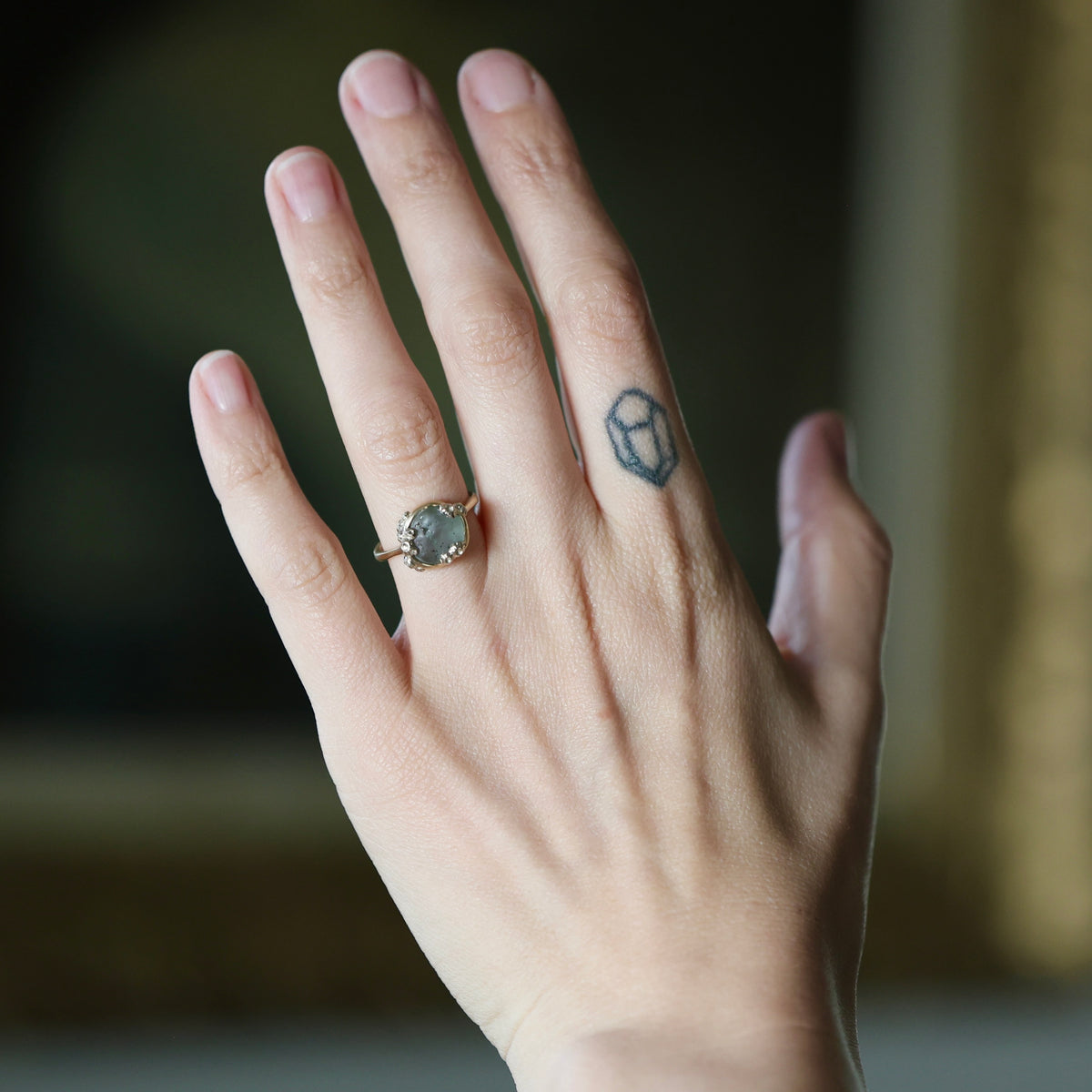 A close-up of a person's hand wearing a gold ring with a round gray stone, with a small geometric tattoo on one of the fingers, against a blurred dark background.