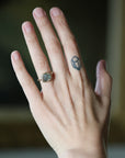 A close-up of a person's hand wearing a gold ring with a round gray stone, with a small geometric tattoo on one of the fingers, against a blurred dark background.