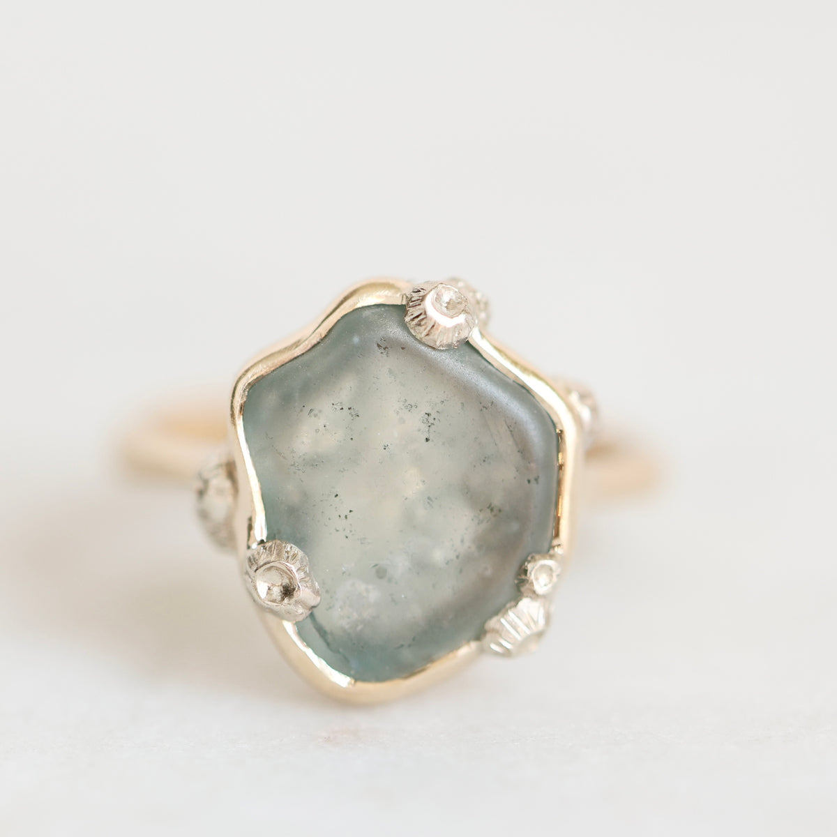 A close-up shot of a raw, shield-shaped light blue-green gemstone in a gold and silver floral bezel setting, on a plain white background.