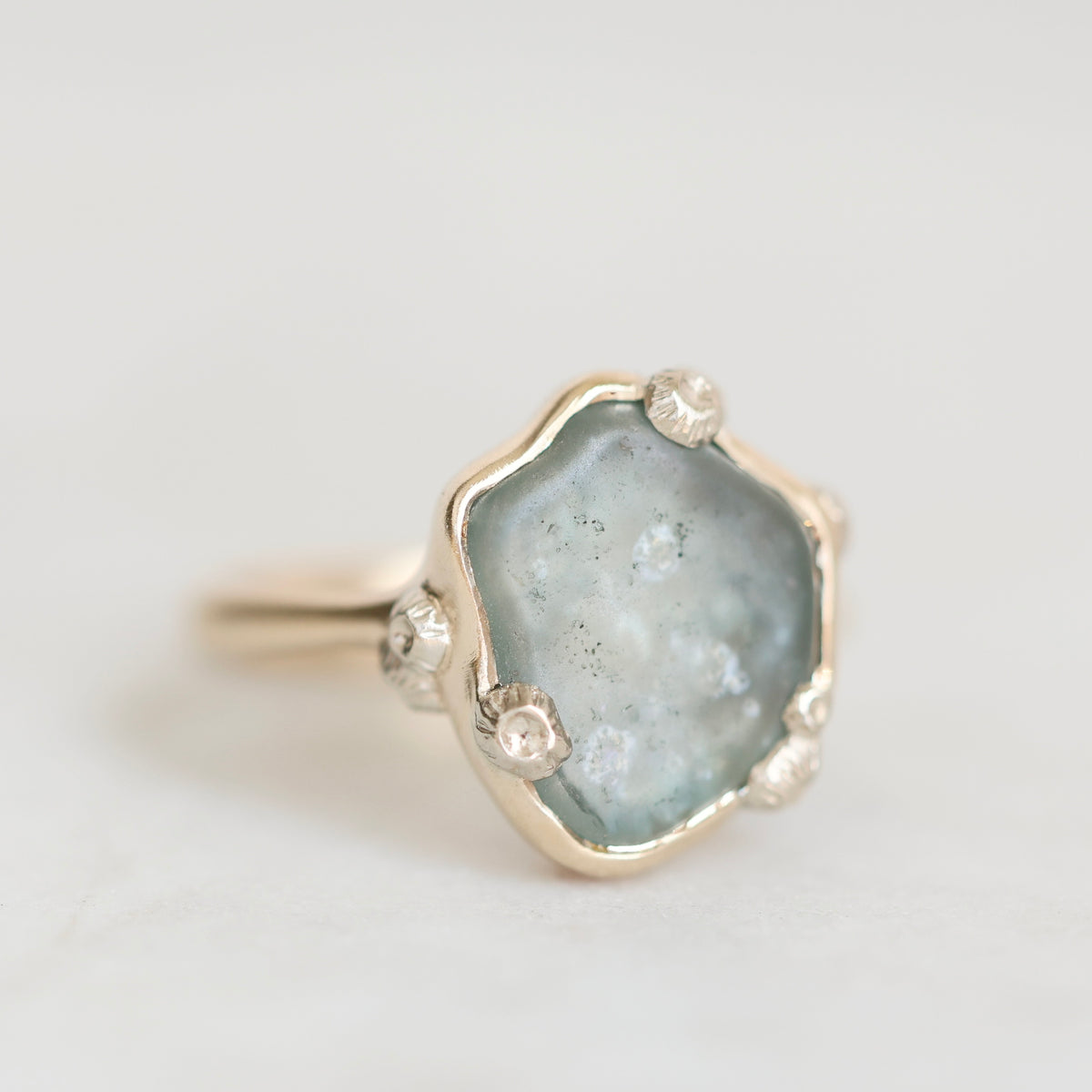 A gold ring featuring a large, irregular-shaped light blue-gray druzy stone with tiny inclusions, accented by three small clear crystals along the band, set against a clean white background.