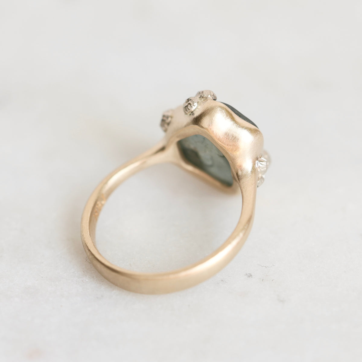 A gold ring with a textured, organic-shaped bezel holding a dark green stone, accented with small clear crystals on the sides, photographed on a light gray surface.
