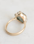 A gold ring with a textured, organic-shaped bezel holding a dark green stone, accented with small clear crystals on the sides, photographed on a light gray surface.