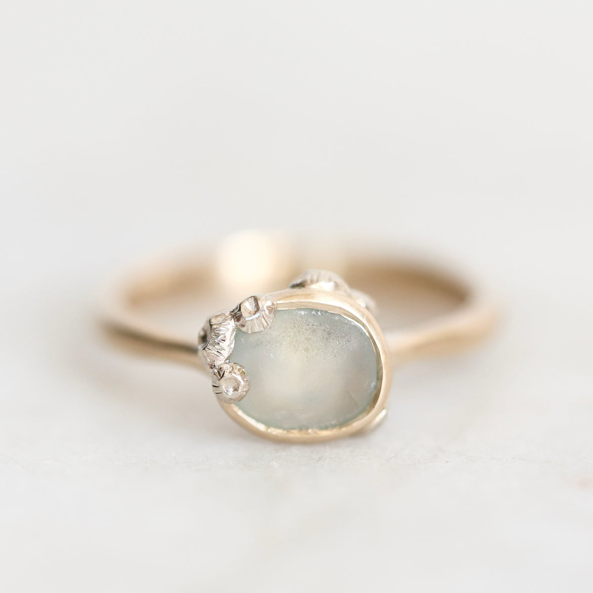 A gold ring with a large, irregular-shaped pale blue-gray stone and small silver accents on the band, photographed on a light gray background.