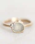 A gold ring with a large, irregular-shaped pale blue-gray stone and small silver accents on the band, photographed on a light gray background.