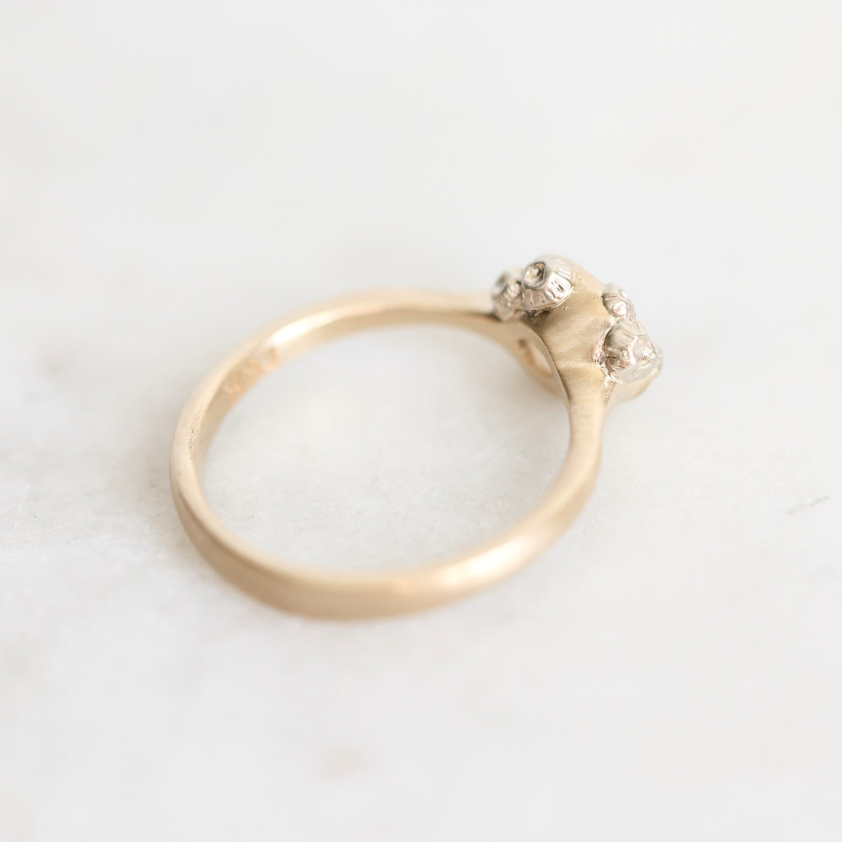 A close-up shot of a gold ring with a textured band and a cluster of small clear gemstones on the setting, resting on a light marble surface.