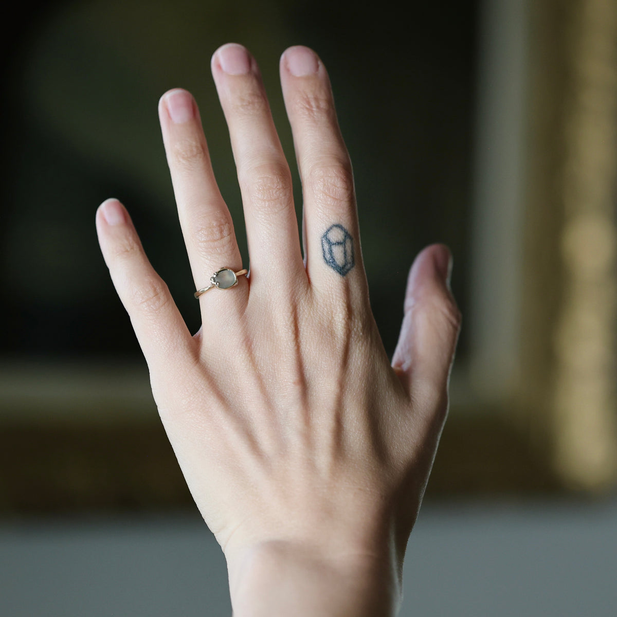 A close-up of a person's hand wearing a delicate gold ring with a round, pale gray stone on the ring finger, alongside a small geometric tattoo on the middle finger, set against a softly blurred dark background.