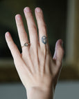 A close-up of a person's hand wearing a delicate gold ring with a round, pale gray stone on the ring finger, alongside a small geometric tattoo on the middle finger, set against a softly blurred dark background.