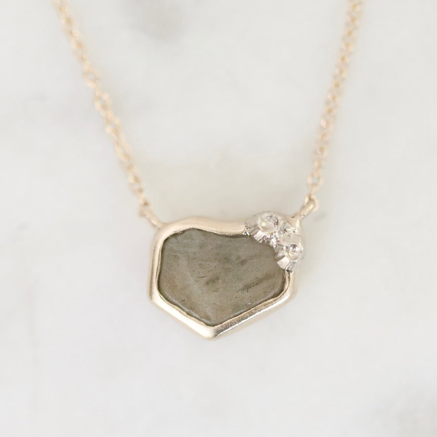 A delicate gold chain necklace featuring an irregular, raw stone pendant with a small cluster of clear crystals at the top right, displayed on a light marble surface.