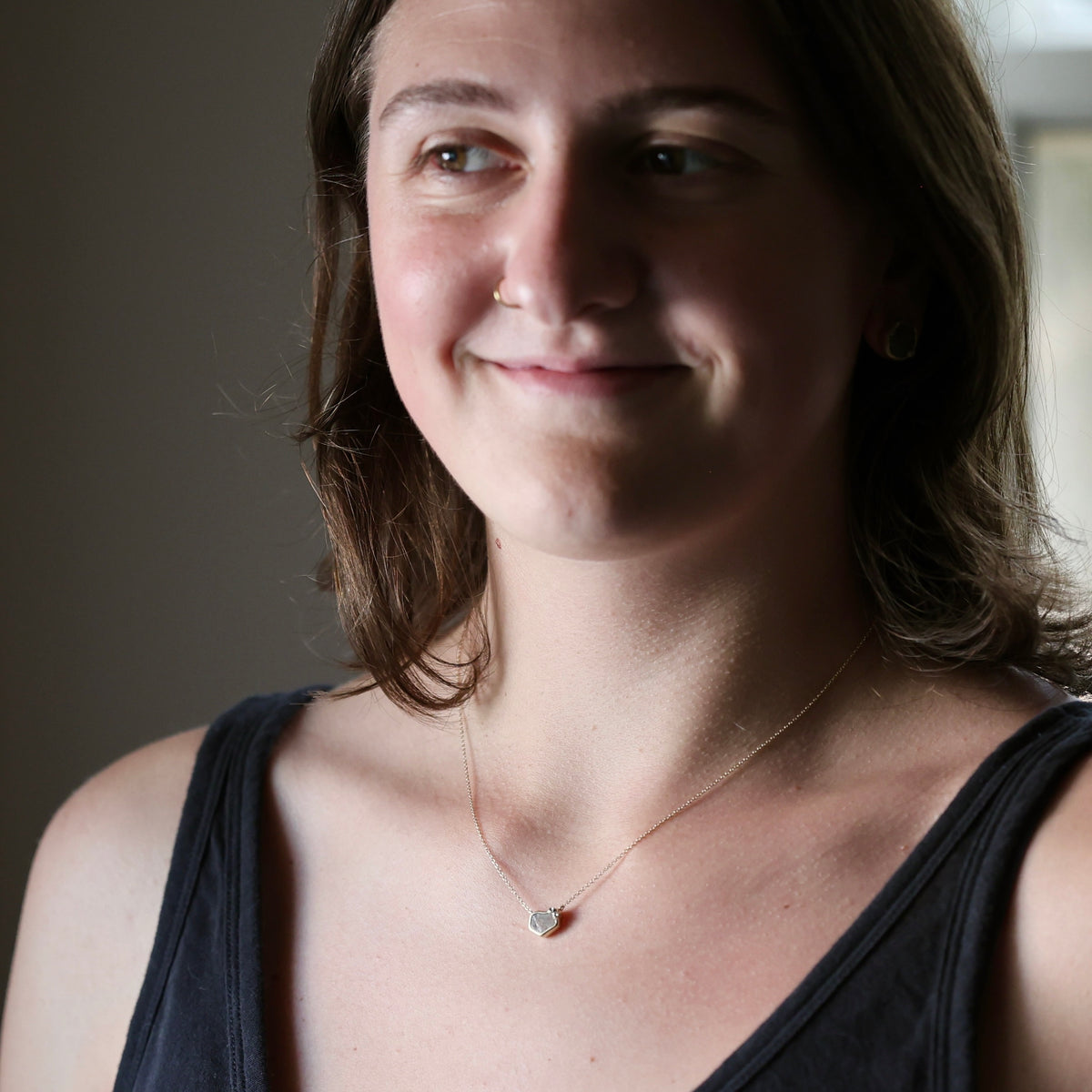 A close-up portrait of a woman wearing a delicate gold chain necklace with a small, round, light-colored stone pendant, set against a soft-focus indoor background.