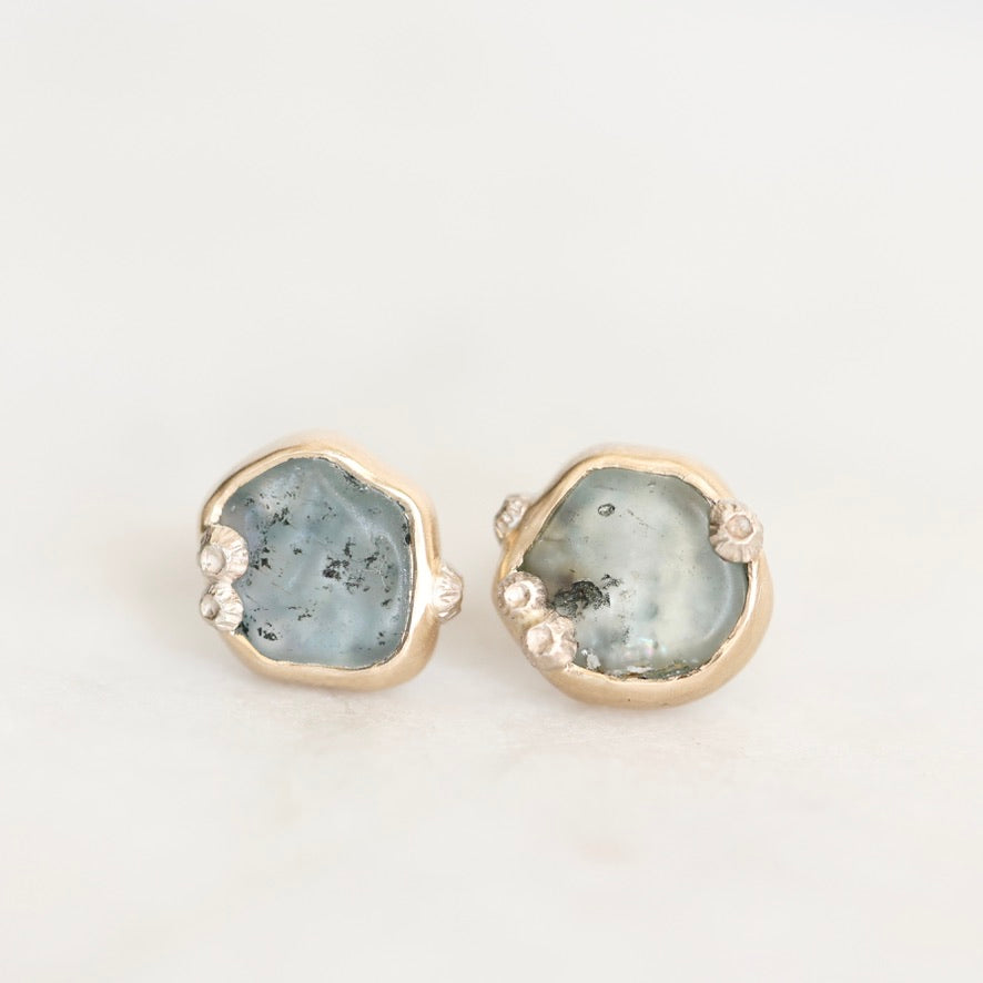 A pair of gold stud earrings featuring irregular, polished light blue  stone slices with tiny clear accent stones along the edges, set against a plain white background.