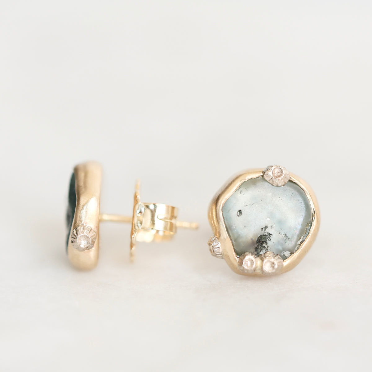 A pair of gold stud earrings on a white background, one showing the front with a polished irregular-shaped pale blue-gray stone and small clear accent stones, and the other showing the post and backing.