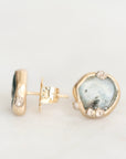 A pair of gold stud earrings on a white background, one showing the front with a polished irregular-shaped pale blue-gray stone and small clear accent stones, and the other showing the post and backing.