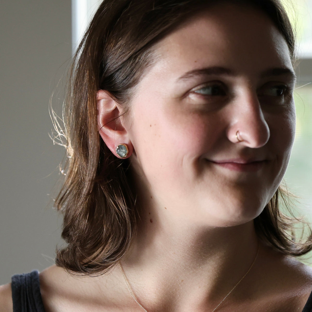 A close-up portrait of a woman with brown hair wearing a small round stud earring with a greenish-gray stone, shown in profile against a softly lit indoor background.
