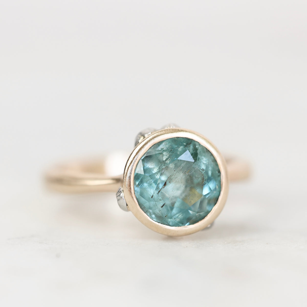 Blue green bezel set gemstone ring with natural inclusions against a plain background. 