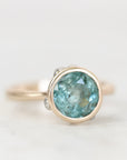 Blue green bezel set gemstone ring with natural inclusions against a plain background. 