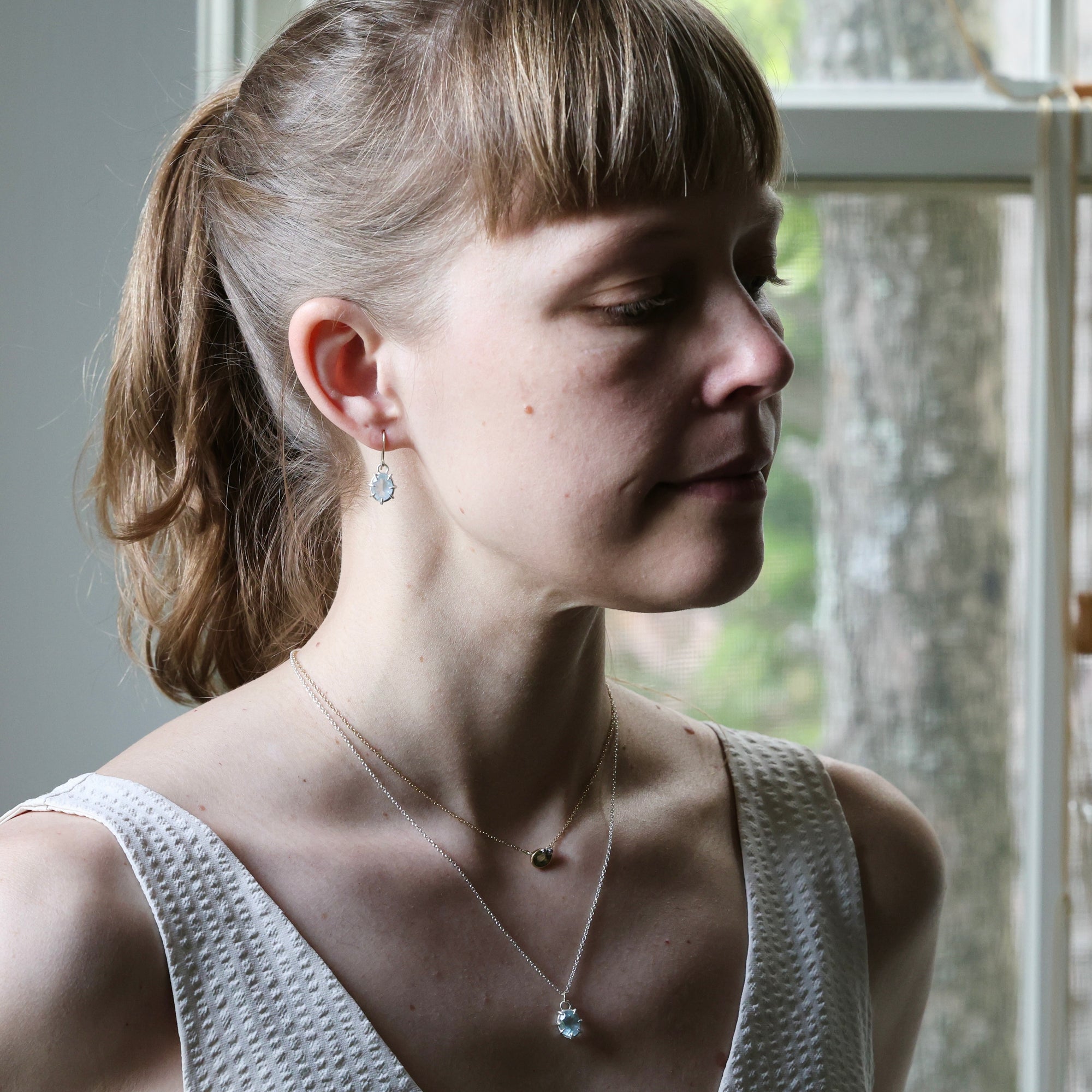 Woman wearing a necklace with a pendant, standing in front of a window.
