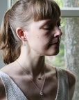 Woman wearing a necklace with a pendant, standing in front of a window.